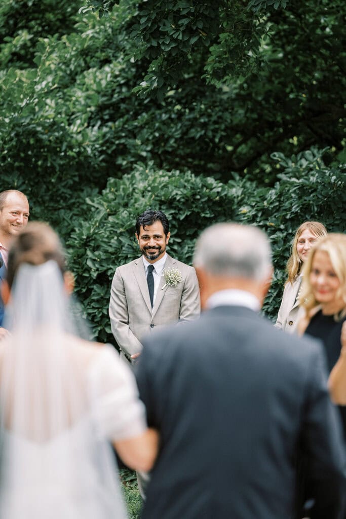 Groom smiling as he sees the bride walk down the aisle during an outdoor wedding at Tyler Arboretum in Philadelphia, surrounded by lush green trees.