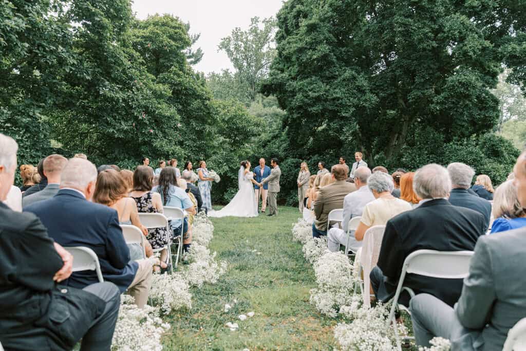 Wide ceremony view of a wedding at Tyler Arboretum in Philadelphia with guests seated along aisles lined with baby’s breath floral arrangements as the couple exchanges vows.