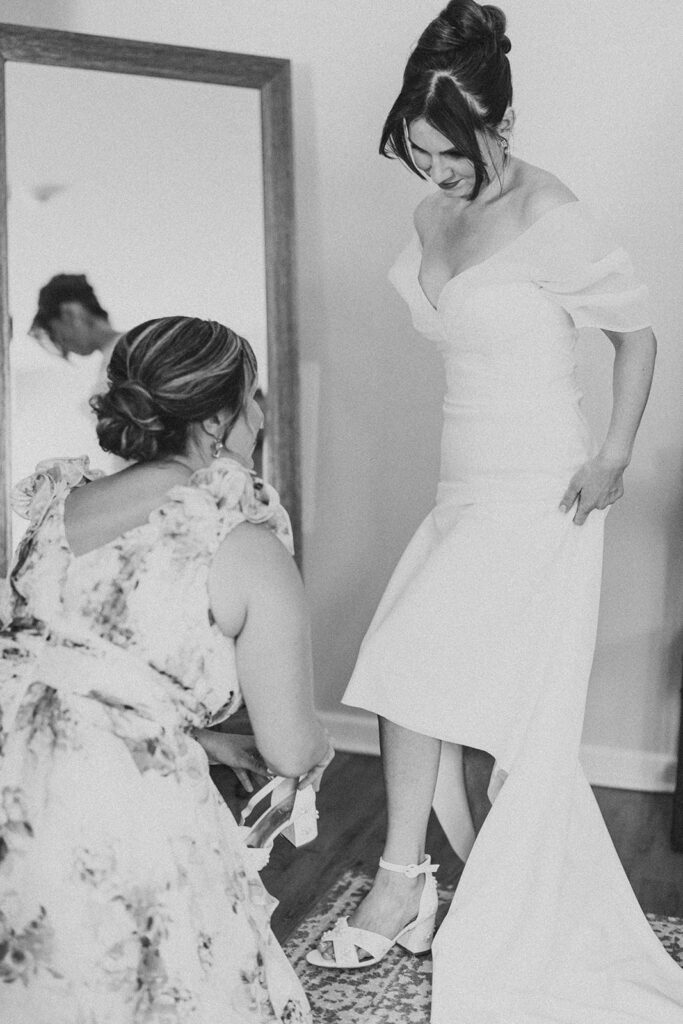 Black and white candid of the bride getting ready for her wedding at Tyler Arboretum in Philadelphia, wearing an off-the-shoulder gown as a bridesmaid in a floral dress helps fasten her daisy-appliqué bridal heels.