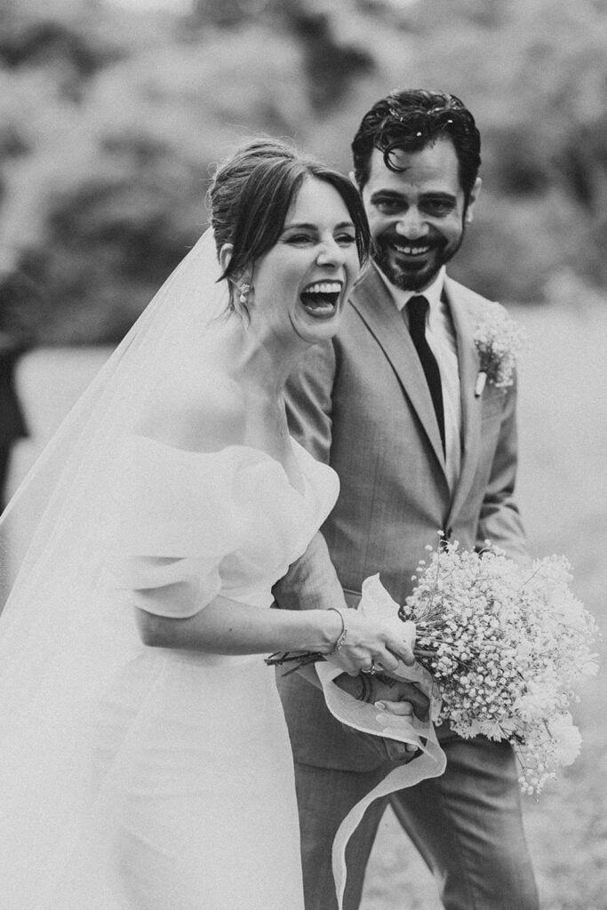 Black and white candid of the bride and groom laughing together after the ceremony, holding baby’s breath wedding florals at Tyler Arboretum in Philadelphia.