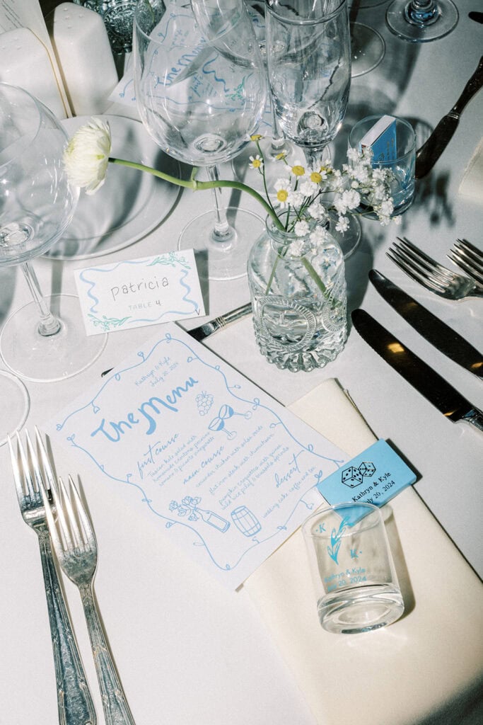Reception table decor at a wedding at Tyler Arboretum in Philadelphia featuring blue illustrated menus, champagne flutes, glass bud vase with chamomile and baby’s breath stems, custom matchbox favors, and handwritten place cards.