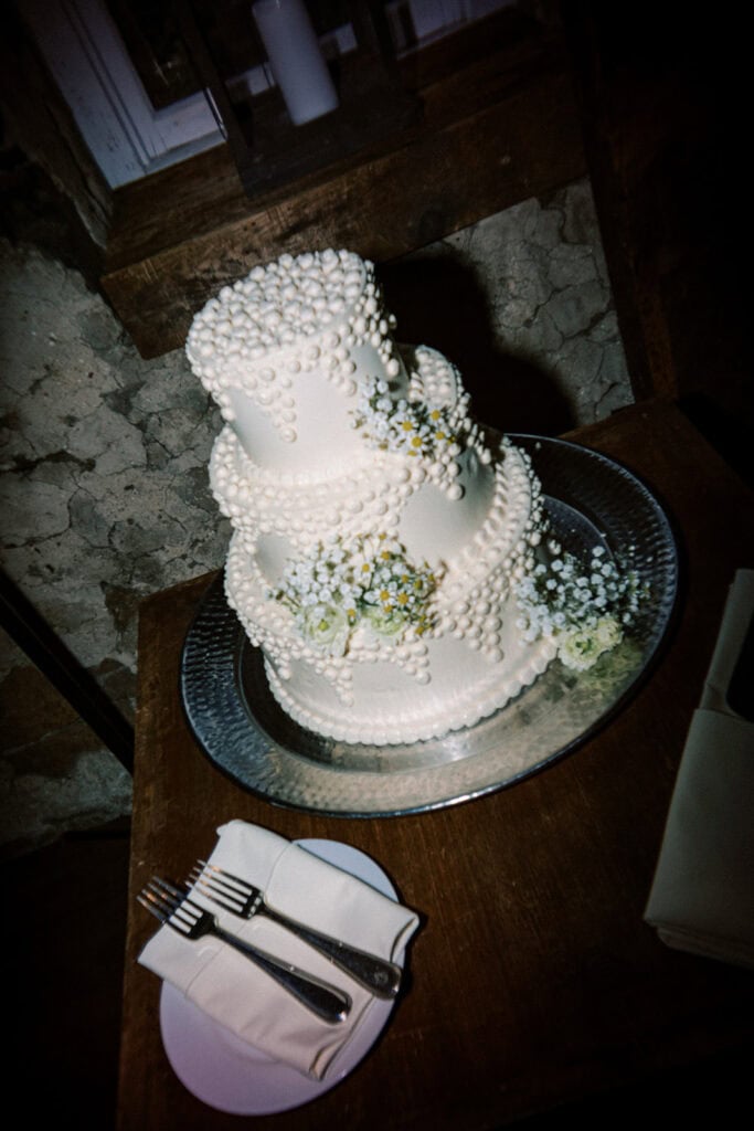 Three-tier white wedding cake decorated with pearl-like piped details and clusters of baby’s breath and chamomile flowers, photographed indoors at a wedding at Tyler Arboretum in Philadelphia.