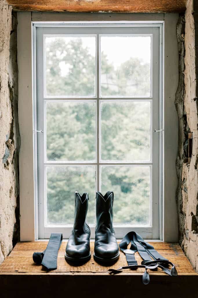 Groom’s black leather boots, navy tie, and suspenders displayed on a rustic wooden windowsill inside a historic building at Tyler Arboretum in Philadelphia.