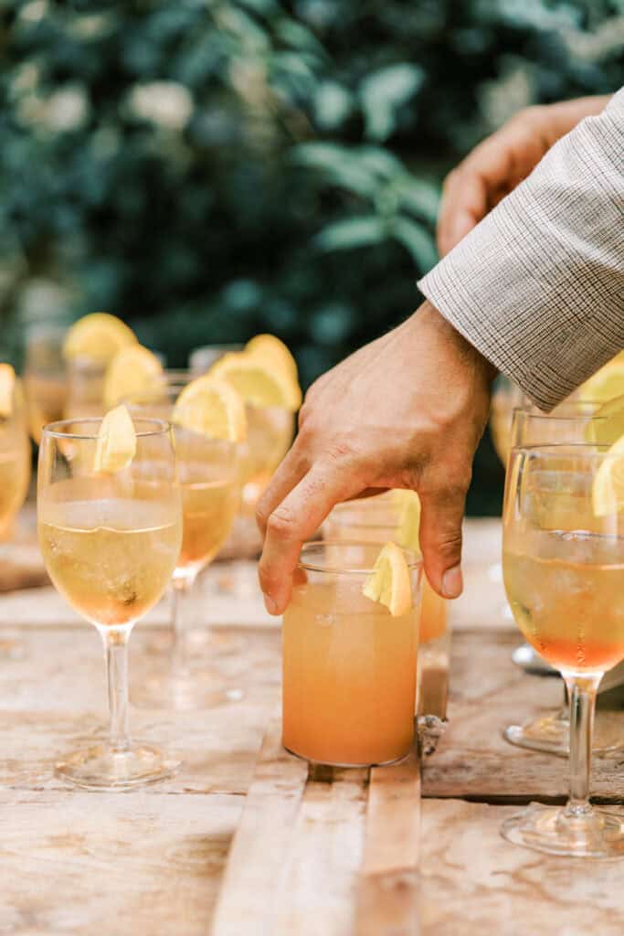 Close-up of signature cocktails served during cocktail hour at a wedding at Tyler Arboretum in Philadelphia, showing iced orange spritz drinks garnished with citrus wedges as a guest reaches for a glass.