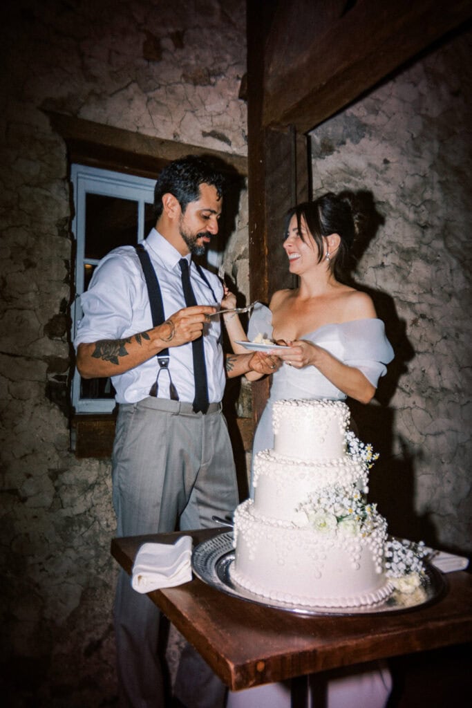 Bride and groom cutting their wedding cake during their reception at Tyler Arboretum in Philadelphia, smiling together beside a three-tier white cake decorated with piped pearl detailing, baby’s breath, and chamomile flowers against the rustic stone interior.