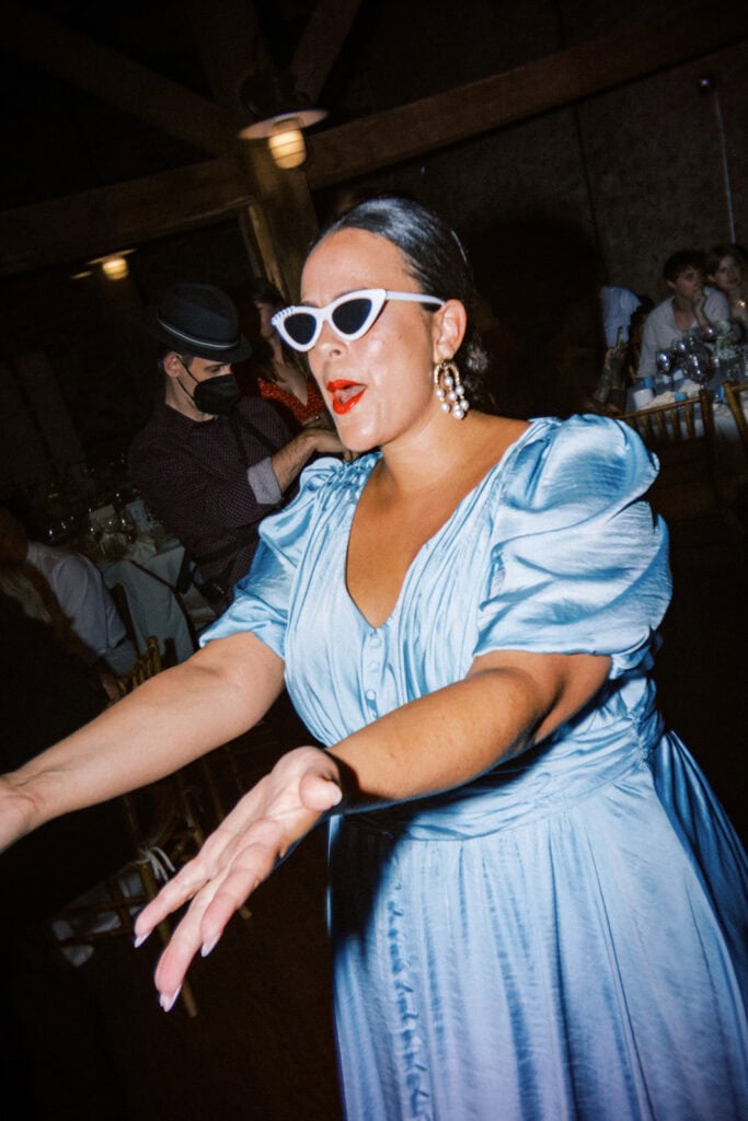 Wedding guest dancing during a reception at Tyler Arboretum in Philadelphia wearing white retro cat-eye sunglasses, pearl statement earrings, and a light blue puff-sleeve satin dress, captured with a flash film aesthetic.