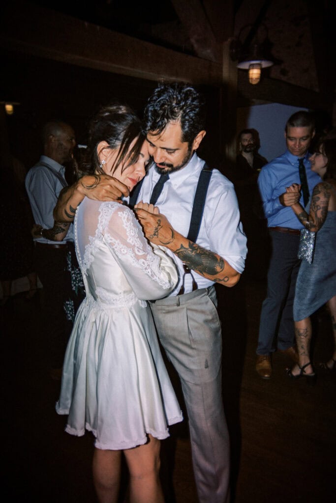 Bride and groom sharing an intimate slow dance at their wedding reception at Tyler Arboretum in Philadelphia, with the bride wearing a vintage-inspired short lace reception dress and the groom in suspenders and a black tie, surrounded by guests on the dance floor.
