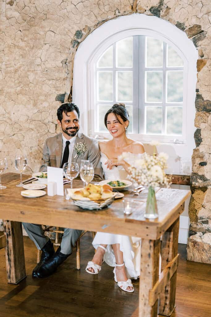 Bride and groom seated at their wooden sweetheart table during a wedding at Tyler Arboretum in Philadelphia, laughing together in front of a rustic stone wall and arched window, with baby’s breath centerpiece and plated dinner.