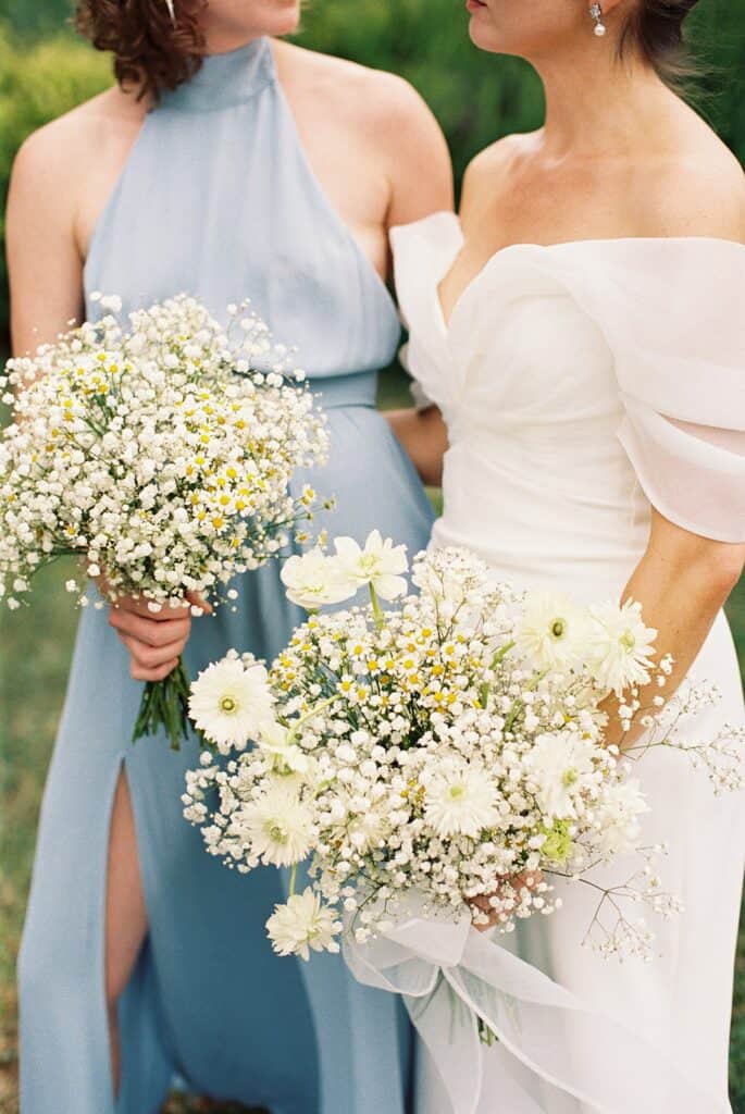 Detail of the bride and bridesmaid holding soft, airy bouquets of white daisies, chamomile, and baby’s breath in blue gowns during a wedding at Tyler Arboretum in Philadelphia.