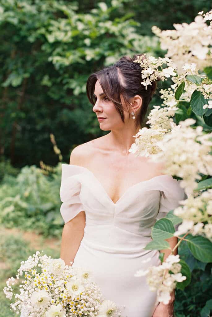 Bride standing beside white hydrangea blooms holding a loose, garden-style bouquet of white daisies, chamomile, and baby’s breath during her wedding at Tyler Arboretum in Philadelphia.