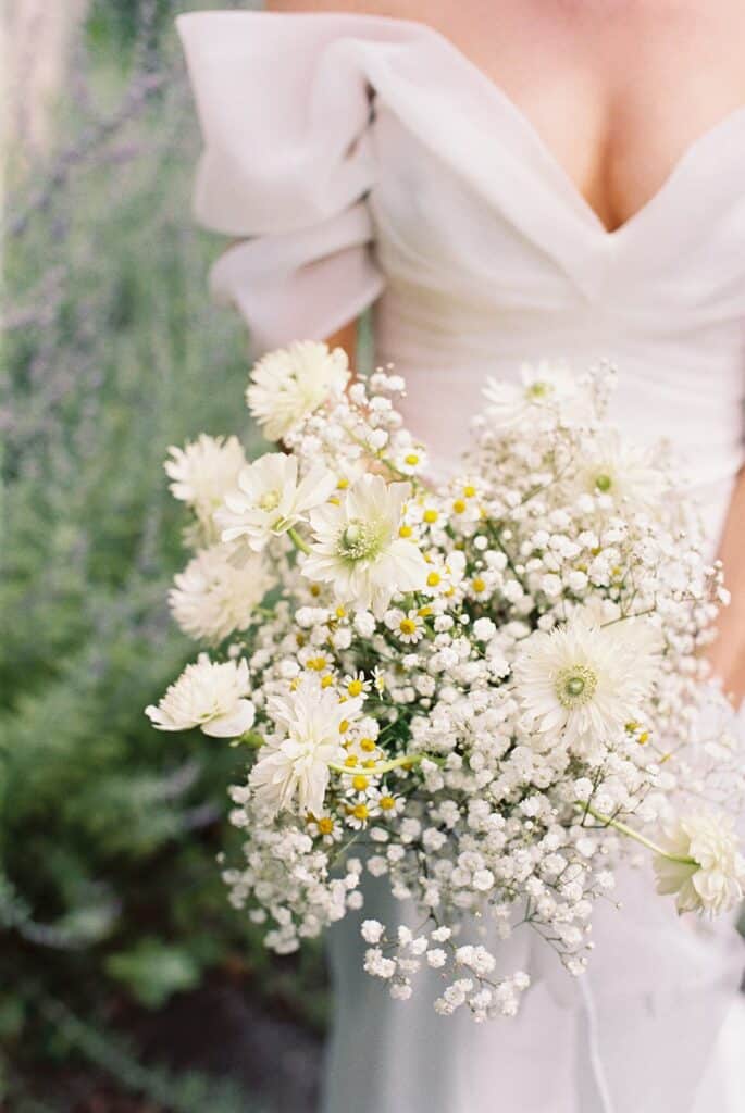Close-up of the bride’s bouquet featuring white daisies, chamomile, and baby’s breath against her off-the-shoulder gown during a wedding at Tyler Arboretum in Philadelphia.