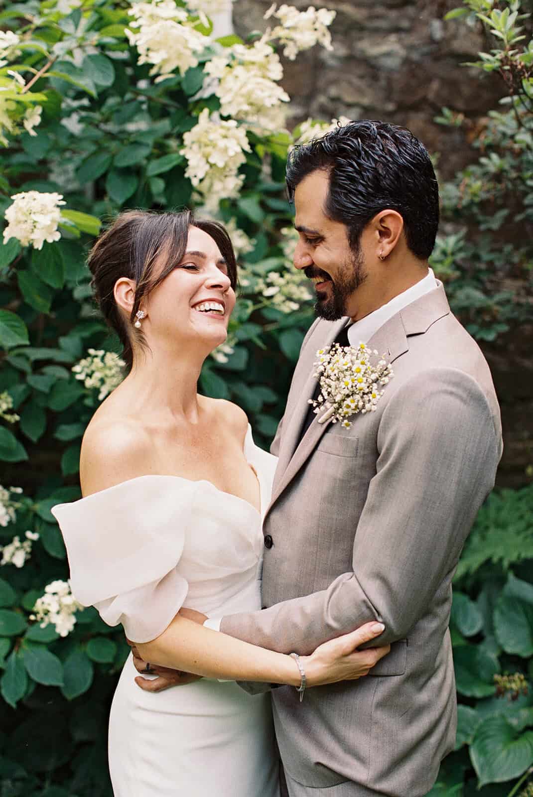 Bride and groom laughing together in front of blooming white hydrangeas, holding baby’s breath and chamomile wedding florals at Tyler Arboretum in Philadelphia.