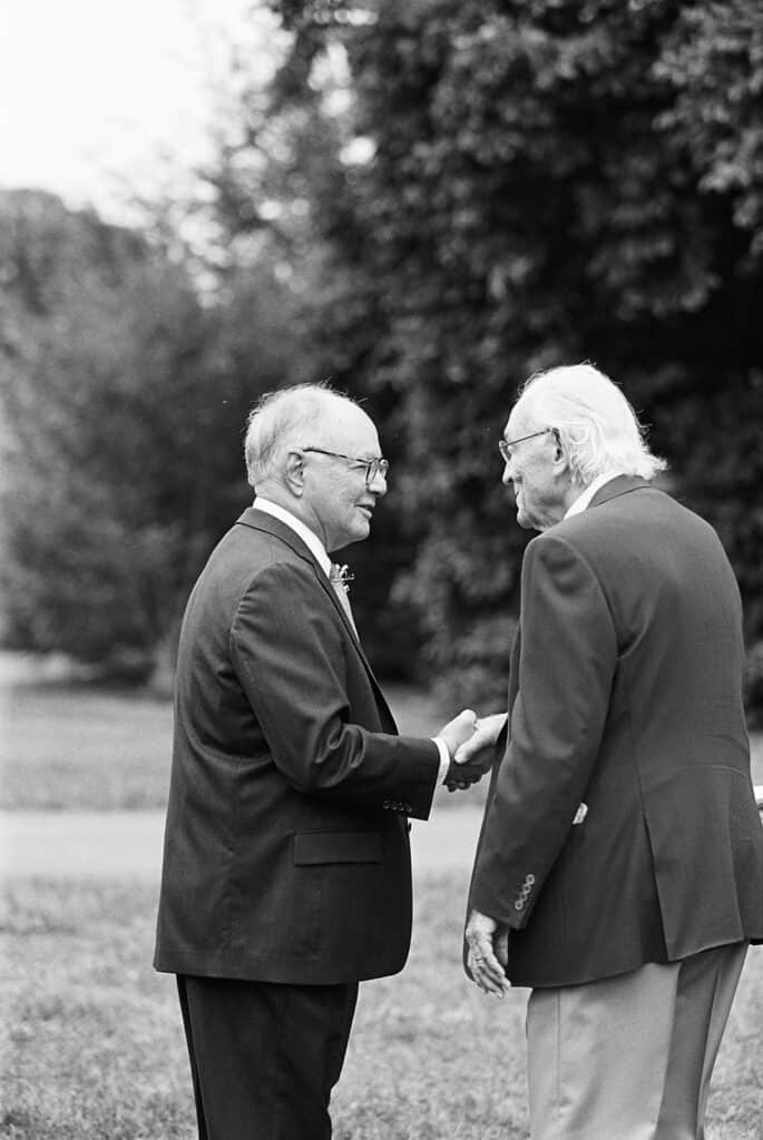 Black and white candid of two older wedding guests shaking hands and greeting one another outdoors at Tyler Arboretum in Philadelphia before the wedding ceremony.