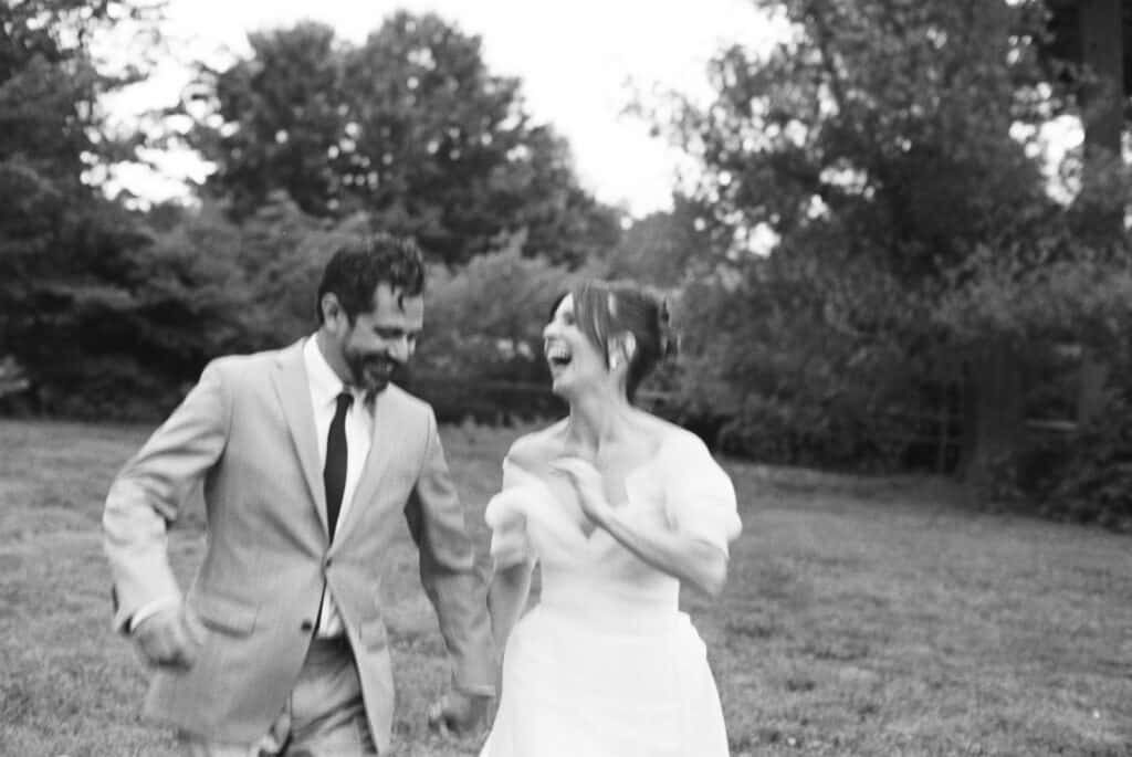 Black and white film photo of the bride and groom running and laughing together on the lawn at Tyler Arboretum in Philadelphia during their wedding day, surrounded by tall summer trees.