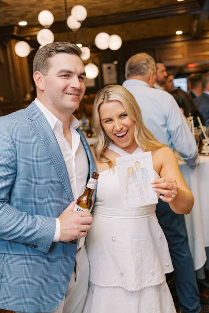 Brenna and Fran smiling together while holding their live watercolor portrait created during their rehearsal dinner at Autograph Brasserie, surrounded by guests and warm ambient light.