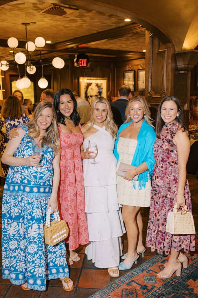 Group of wedding guests posing together inside Autograph Brasserie during Brenna and Fran’s rehearsal dinner, wearing colorful spring dresses and holding drinks in a warmly lit restaurant space.