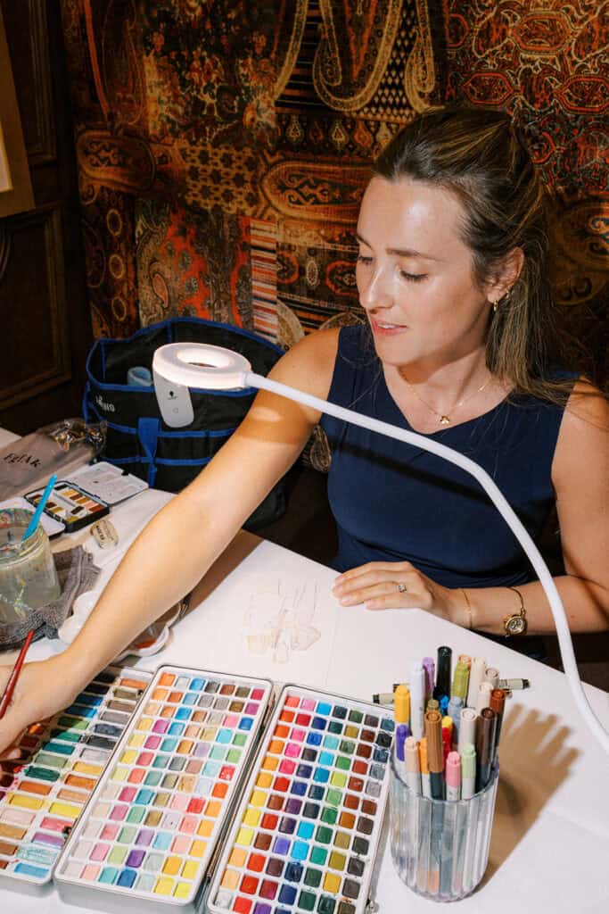 Wedding live painter from Callie Co Portraits painting a watercolor portrait at Autograph Brasserie during Brenna and Fran’s rehearsal dinner, surrounded by art supplies and warm interior lighting.