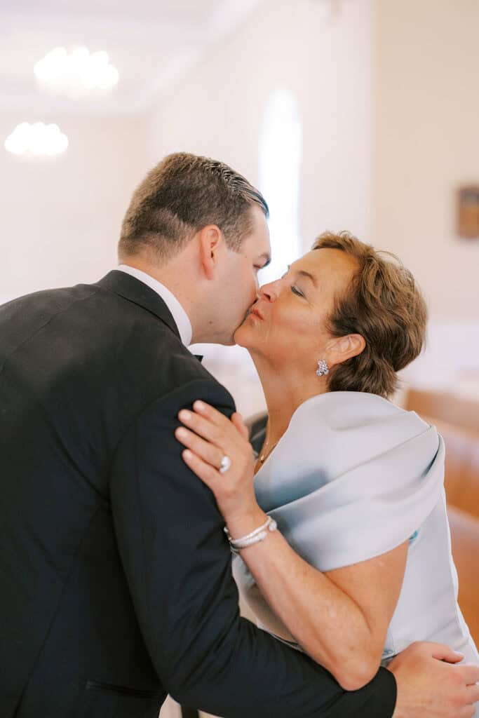 Close-up of the groom in a tux kissing his mom on the cheek inside a church, her arm wrapped around him and a ring visible on her hand.