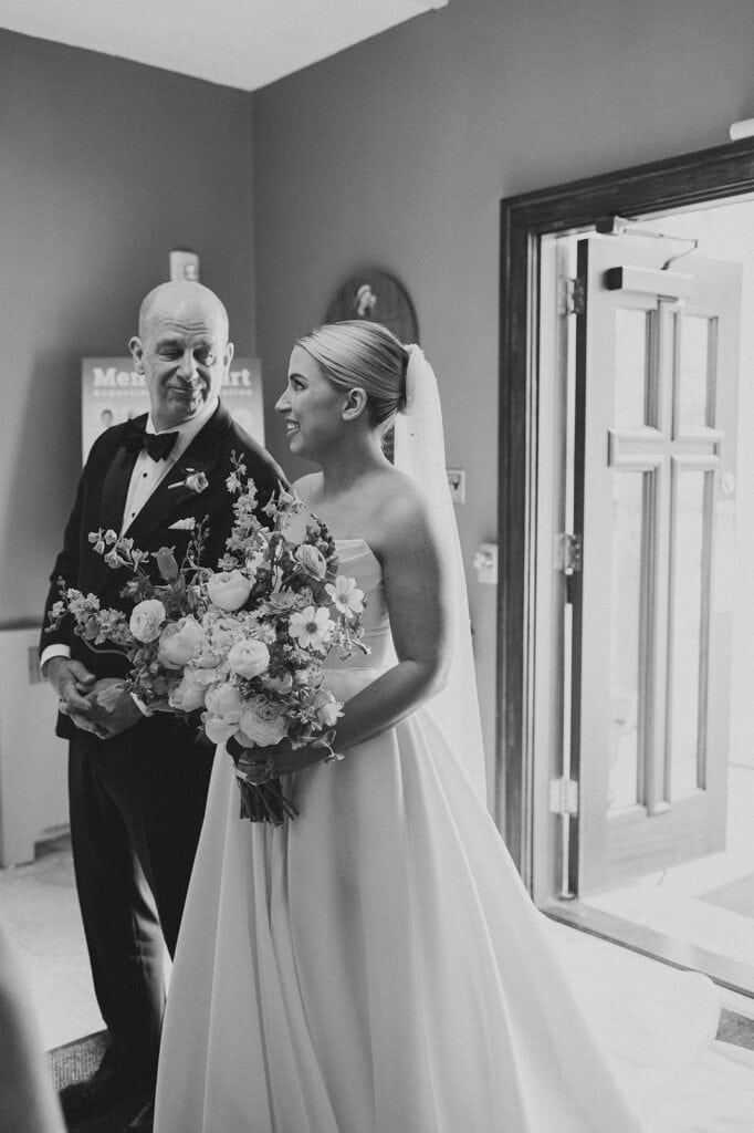 Black and white photo of the bride in a strapless gown and veil holding a bouquet, smiling at an older man in a tux near an open doorway.