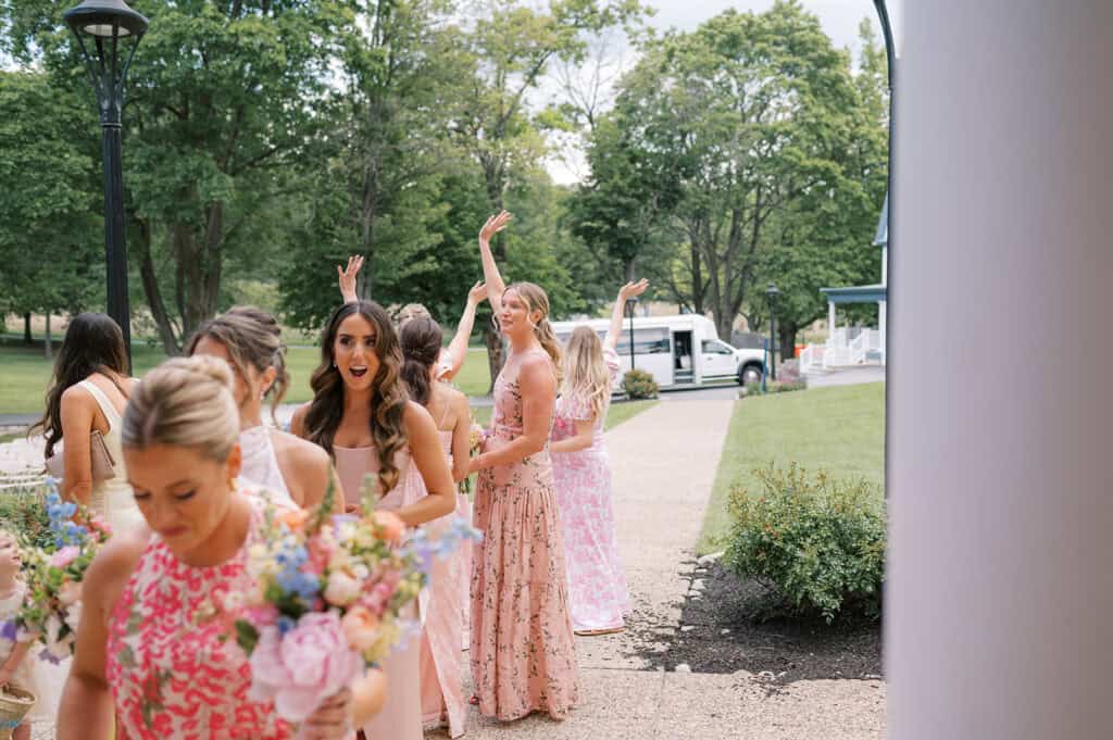Bridesmaids in blush pink and floral dresses arrive outside the ceremony space, holding colorful bouquets while one bridesmaid raises her arms playfully in the background.