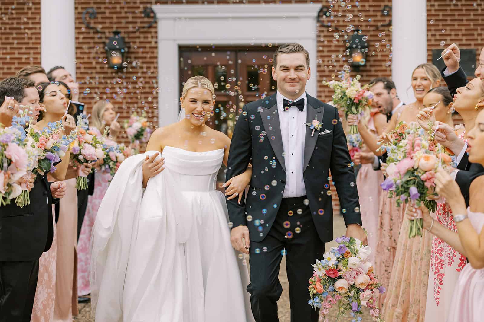 Bride and groom walking hand in hand through a bubble exit outside Our Mother of Good Counsel Chapel, surrounded by wedding party and guests holding colorful floral bouquets.