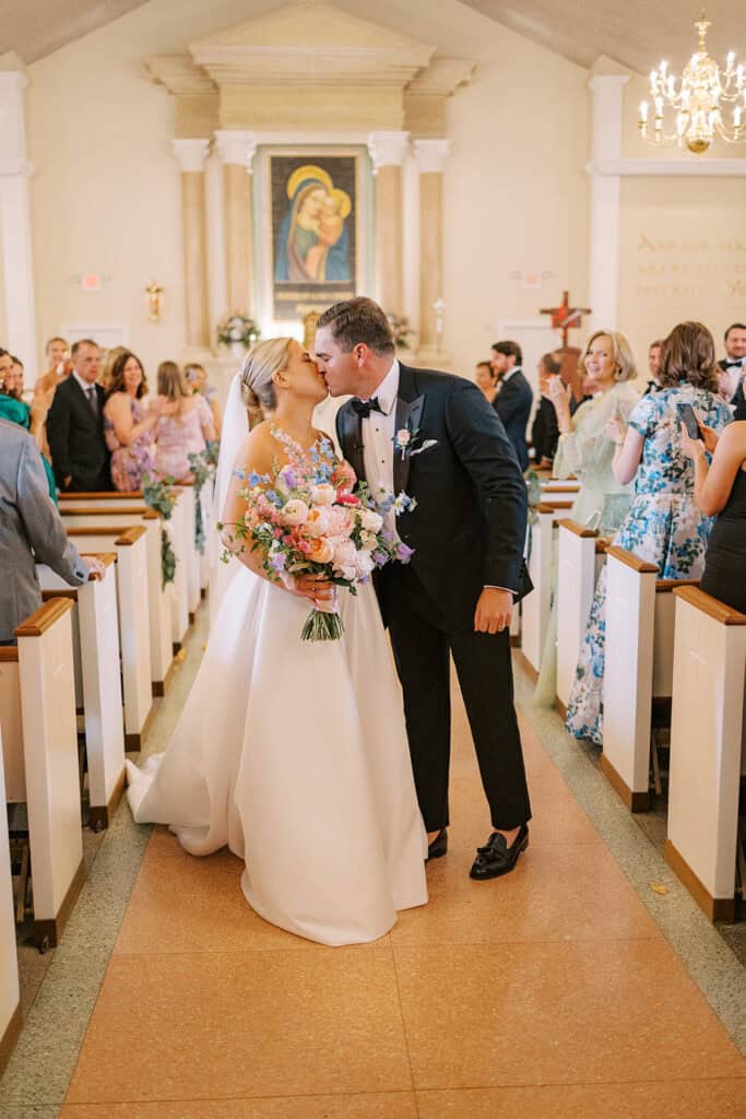 The bride and groom share their first kiss in the church aisle following the ceremony, surrounded by guests standing and applauding from the pews.