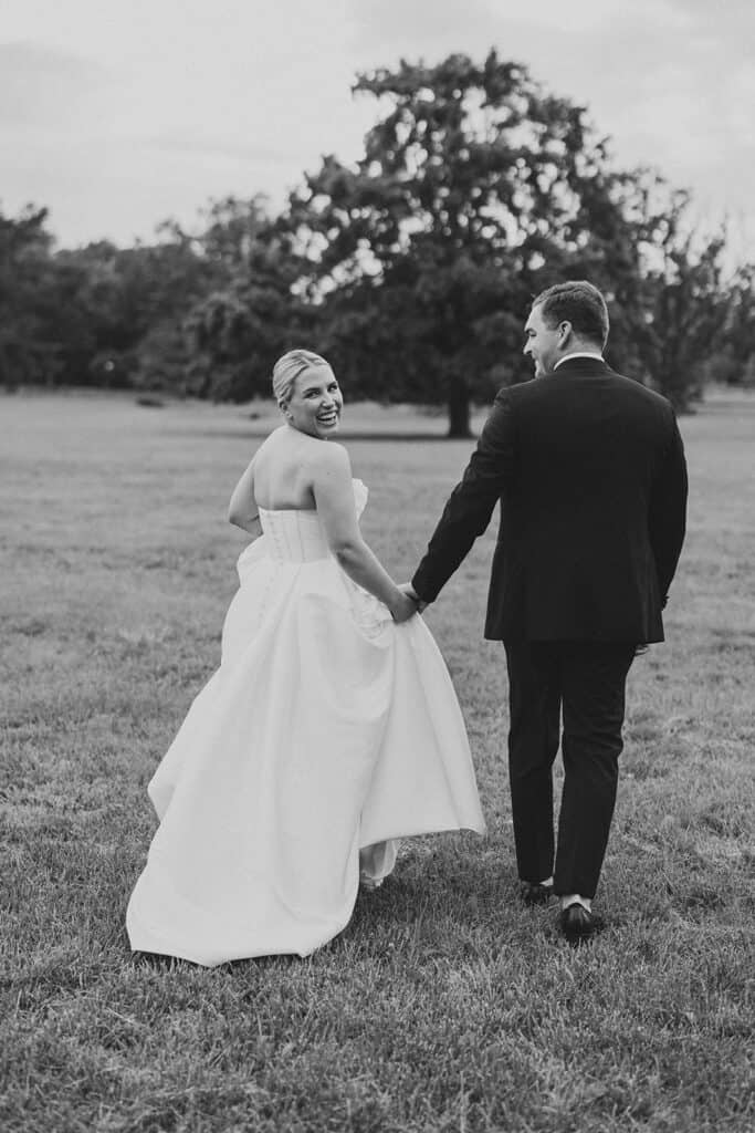 Black and white portrait of the bride and groom walking away together across a grassy field outside The Ivy at Ellis Preserve, with the bride looking back over her shoulder and smiling.