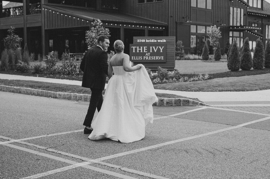 Black and white portrait of the bride and groom walking together outside The Ivy at Ellis Preserve, with the venue sign visible in the background as the bride lifts her wedding gown and the groom looks back toward her.