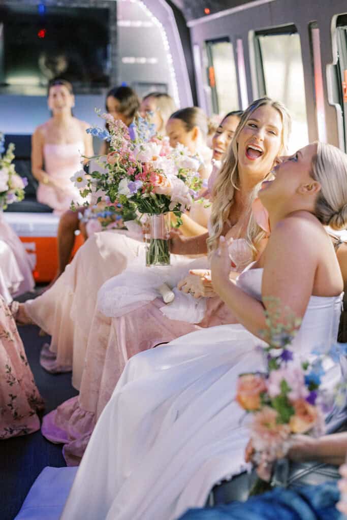 Bride and bridesmaids seated inside a party bus holding bouquets and wearing pink dresses, with the bride seated in the foreground laughing while holding her skirt.