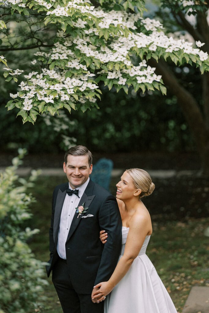 Bride standing behind the groom with her arm around him, laughing during portraits outside The Ivy at Ellis Preserve under white flowering dogwood branches, with a blue garden bench visible in the background.