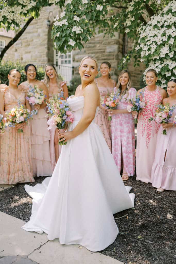 Bride standing in the foreground holding a colorful bouquet while bridesmaids stand behind her beneath blooming white dogwood trees, all wearing pink and blush dresses outside The Ivy at Ellis Preserve.