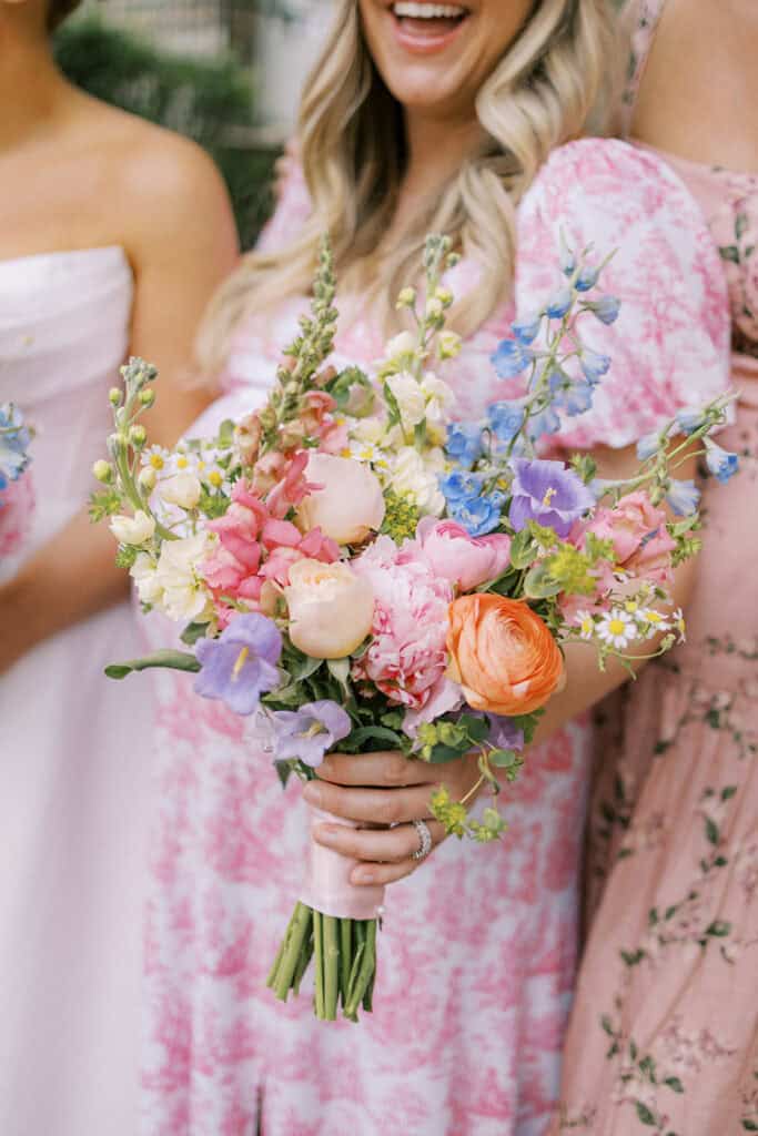 Close-up of a bridesmaid holding a colorful bouquet featuring pink, peach, blue, and white flowers, with a pink patterned bridesmaid dress visible in the background.