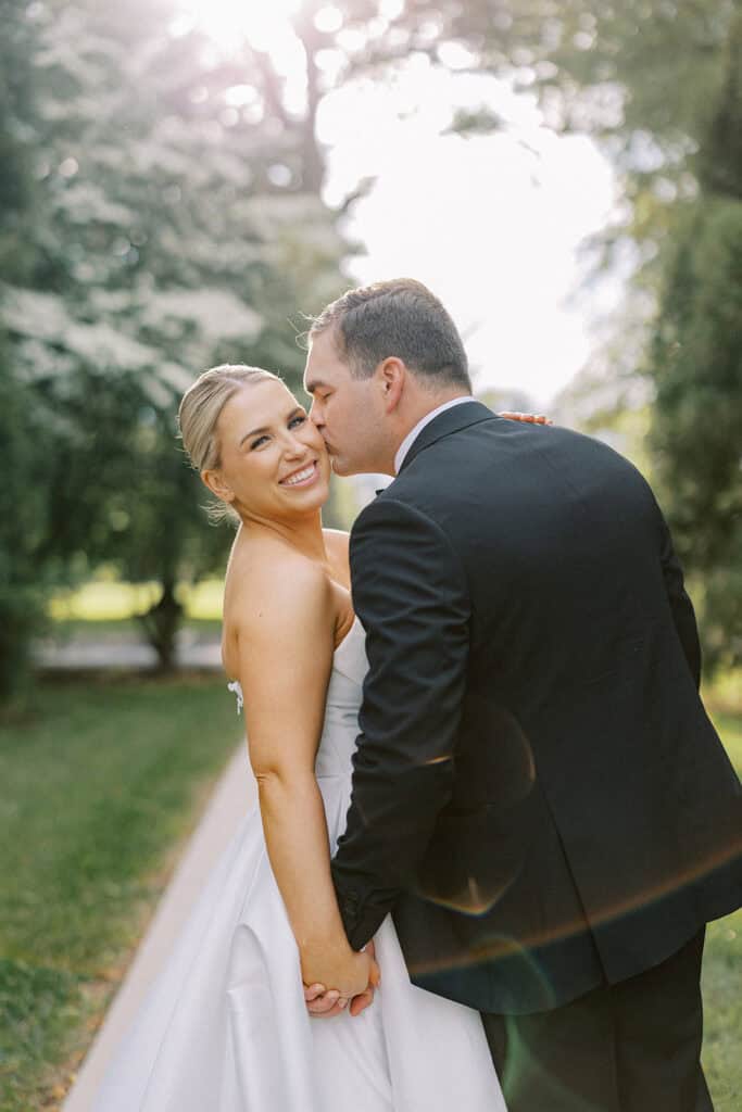 Groom kissing the bride on the cheek while she smiles toward the camera during golden hour portraits outside The Ivy at Ellis Preserve, with backlit trees and natural lens flare.