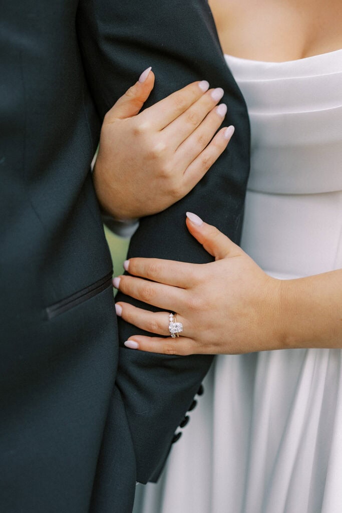 Close-up of the bride’s hands resting on the groom’s arm, highlighting a diamond engagement ring and pale pink manicure during bride and groom portraits outside The Ivy at Ellis Preserve.
