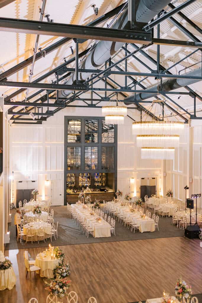 Elevated view of The Ivy at Ellis Preserve ballroom showing long reception tables, round guest tables, floral centerpieces, chandeliers, and exposed beams.