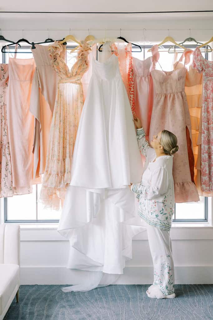 Bride standing in pajamas adjusting her strapless wedding dress displayed among blush bridesmaid dresses during getting ready at The Ivy at Ellis Preserve