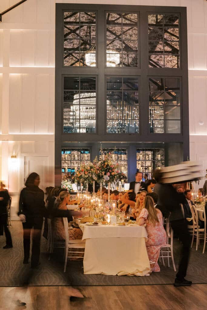 Guests seated at a long reception table during dinner at The Ivy at Ellis Preserve, with tall floral centerpieces, glowing candles, and servers moving through the room.