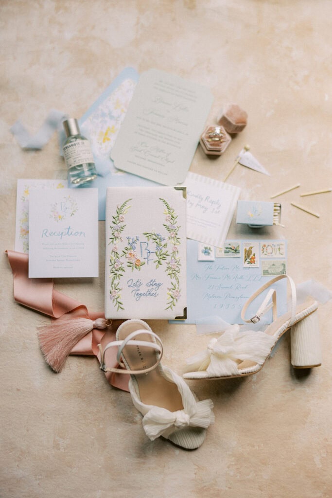 Full wedding invitation suite with pastel blue envelopes, floral paper goods, stamps, ribbon, perfume bottle, shoes, and rings styled for a spring wedding at The Ivy at Ellis Preserve