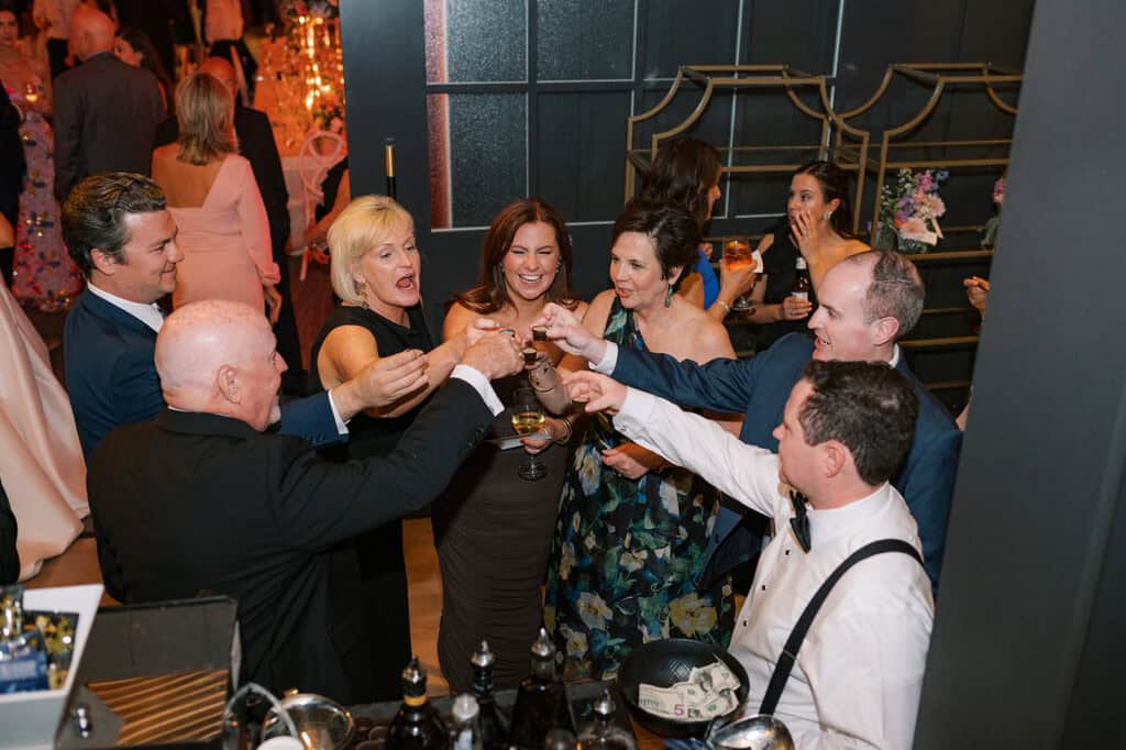 Group of wedding guests gathered around the bar clinking shot glasses and laughing together during the reception at The Ivy at Ellis Preserve.