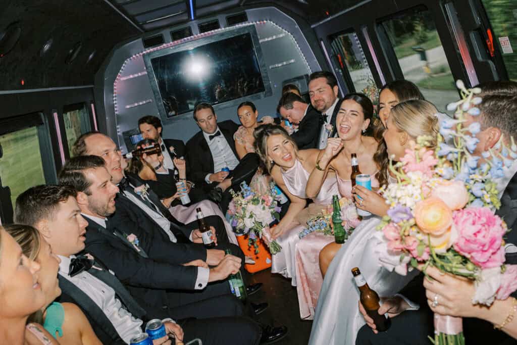 Wedding party laughing and singing together on a shuttle bus while holding bouquets and drinks, traveling from the ceremony to The Ivy at Ellis Preserve reception.
