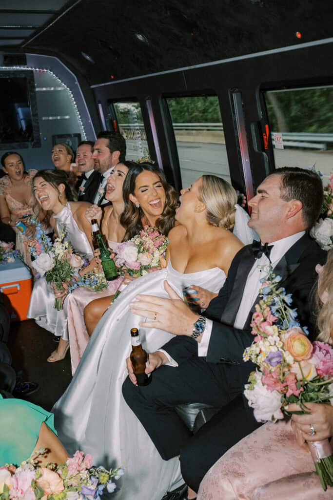 Wedding party laughing and singing together on a shuttle bus while holding bouquets and drinks, traveling from the ceremony to The Ivy at Ellis Preserve reception.