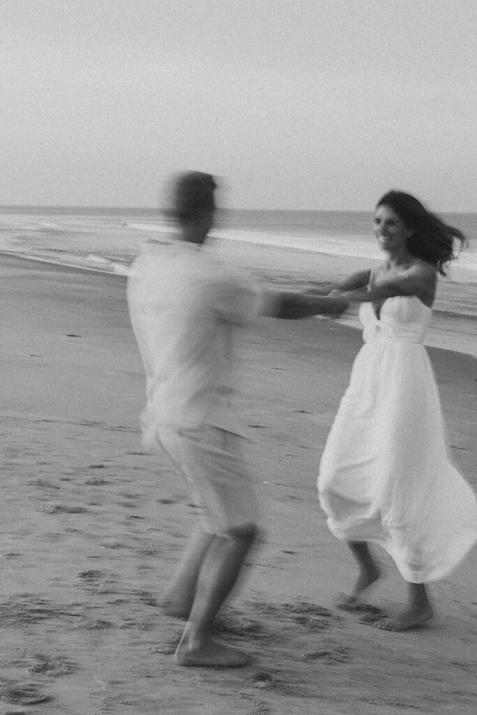 Black and white film photo of a couple demonstrating posing and beach engagement photo ideas by twirling and dancing on Rehoboth Beach during engagement session