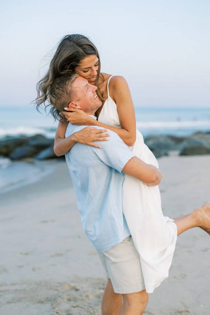 Groom lifting bride along rocky Rehoboth Beach shoreline during engagement photos
