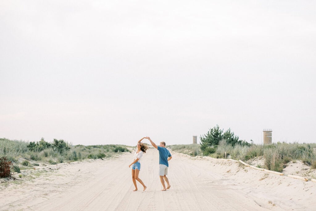 Couple demonstrates posing and beach engagement photo ideas twirling together on a wide sandy beach path during their Rehoboth engagement photoshoot