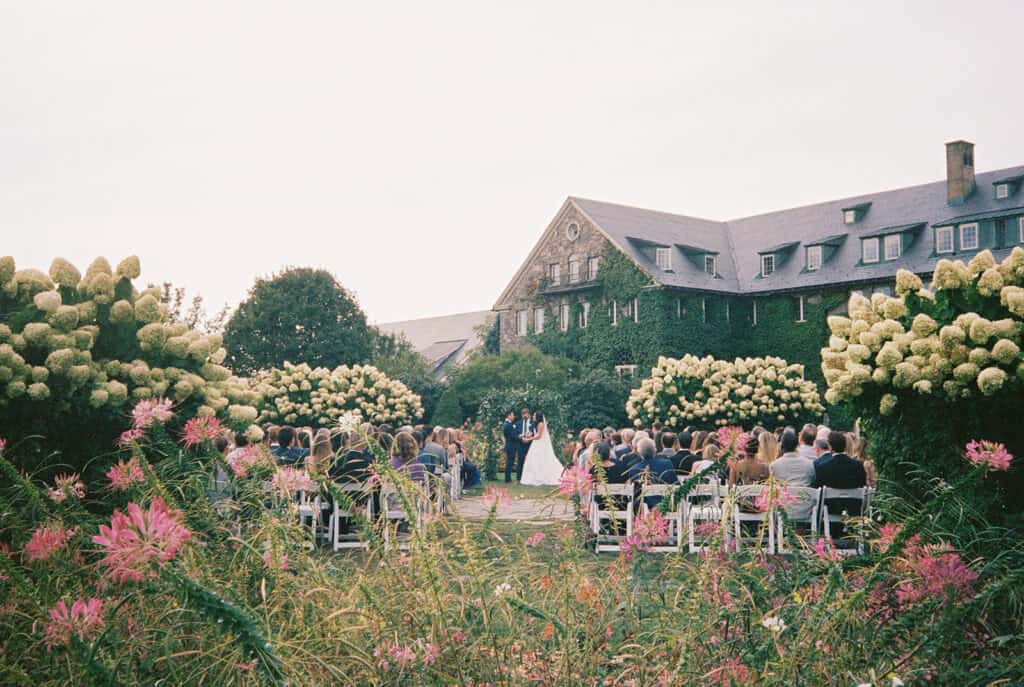 35mm film photo of the outdoor wedding ceremony at Skytop Lodge, captured through wildflowers with blooming hydrangeas and the ivy-covered stone lodge in the background