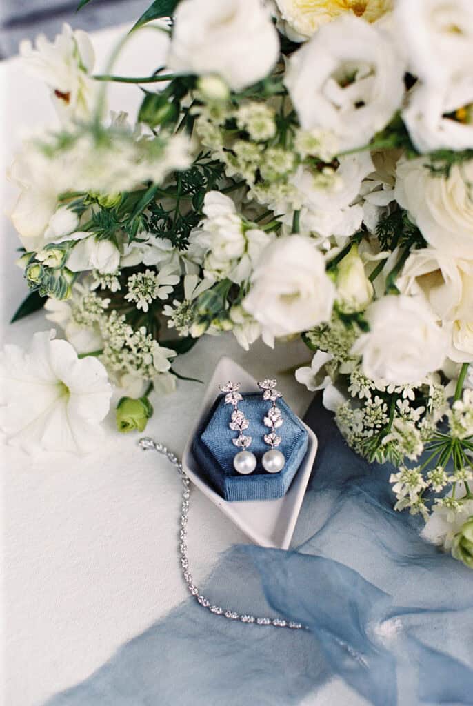 35mm film close-up of pearl and diamond drop earrings in a dusty blue velvet ring box, styled with a diamond tennis bracelet and white garden bridal bouquet for a Skytop Lodge wedding