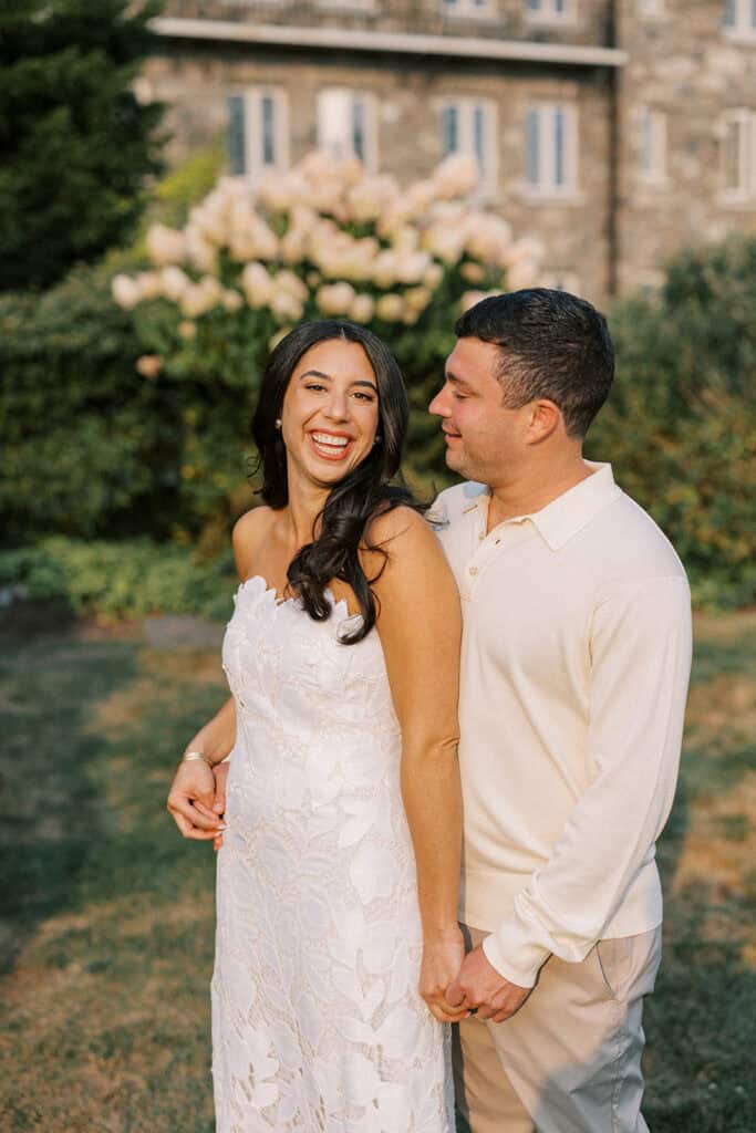 Bride smiling warmly during rehearsal dinner sunset portraits in front of blooming hydrangeas and the stone facade of Skytop Lodge in Skytop, Pennsylvania