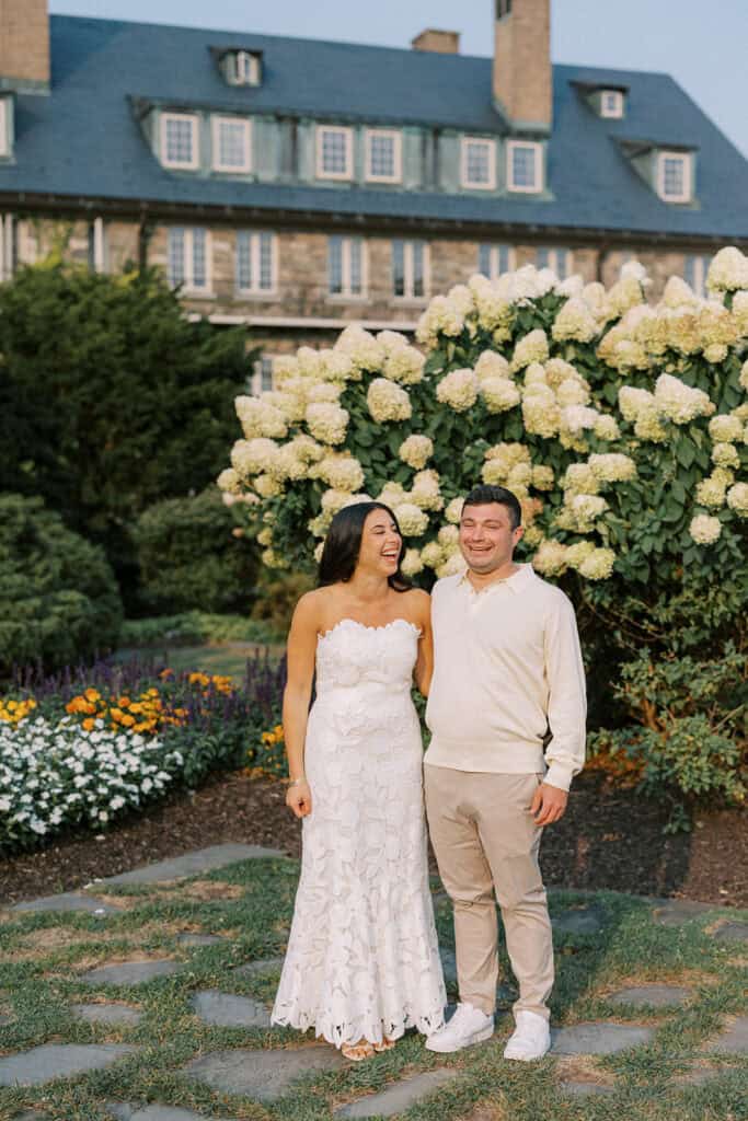 Bride and groom laughing together in rehearsal dinner attire in front of blooming hydrangeas on the grounds of Skytop Lodge during their wedding weekend at Skytop, Pennsylvania
