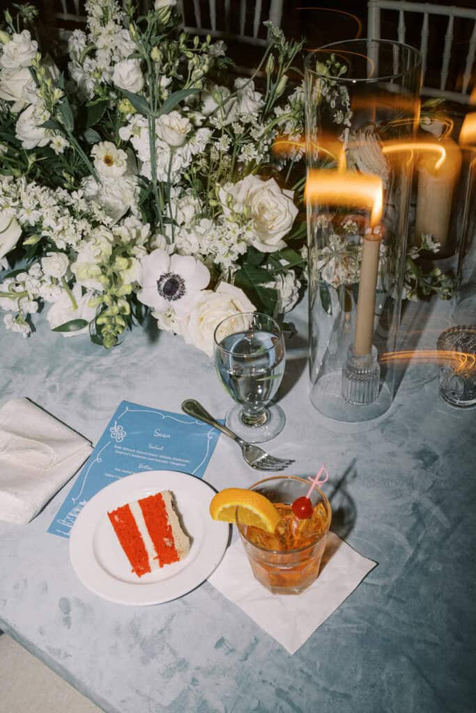 Red velvet wedding cake slice and old fashioned cocktail on a dusty blue linen reception table alongside white floral centerpiece and candlelight at Skytop Lodge