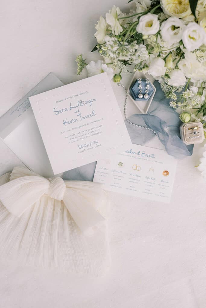 Flat lay of blue and white Skytop Lodge wedding invitation suite with bridal bouquet, diamond earrings, engagement ring, and silk ribbon in Skytop, Pennsylvania